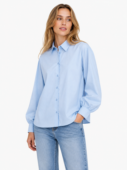 Eine Frau, die die langärmelige, schmal geschnittene Bluse „Antonia Slim“ in Hellblau trägt, kombiniert mit einer hellblauen Jeans, steht vor einem weißen Hintergrund, blickt leicht nach links und hat eine Hand in der Tasche.