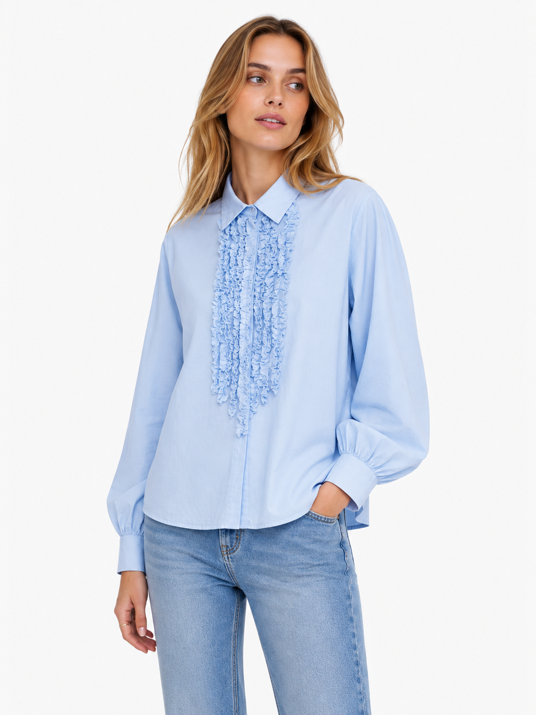 Eine Frau, die die „Antonia Slim Ruffle Light Blue“ trägt – eine Bluse aus einem weichen Baumwoll-Voile-Mix mit eleganten Rüschenverzierungen auf der Vorderseite –, kombiniert mit hellblauen Jeans, steht vor einem schlichten weißen Hintergrund.