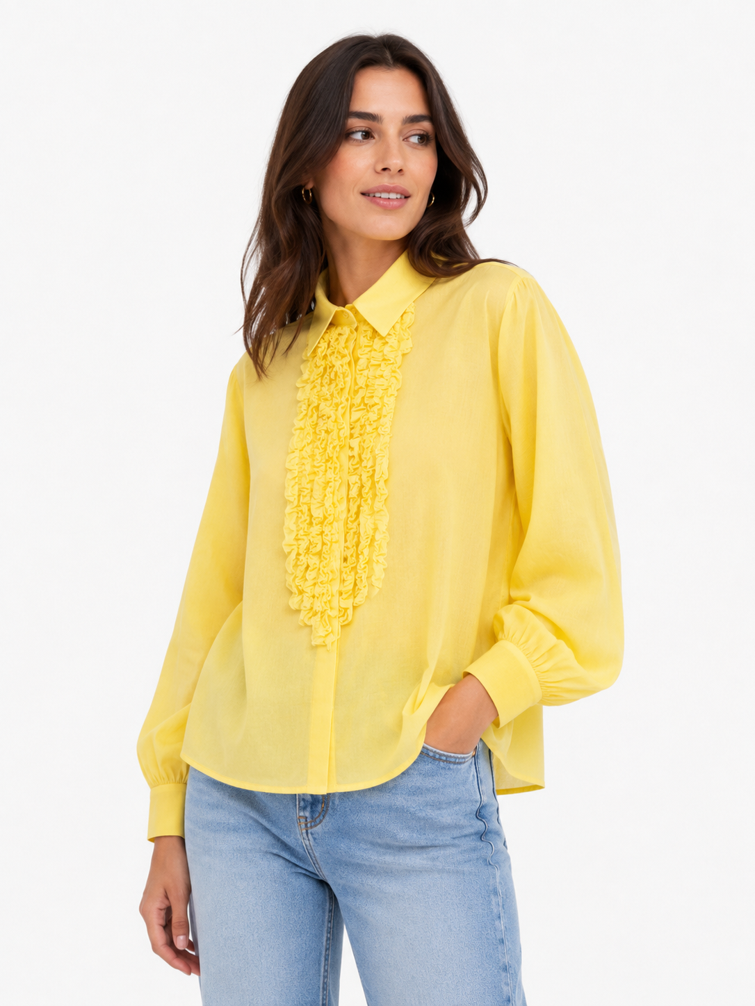 Eine Frau trägt die „Antonia Slim Ruffle Yellow“, eine feminine, schmal geschnittene Bluse aus einem weichen Baumwoll-Voile-Mix in leuchtendem Gelb mit Rüschenverzierungen auf der Vorderseite, dazu eine hellblaue Jeans. Sie steht mit einer Hand in der Tasche und blickt vor einem weißen Hintergrund zur Seite.