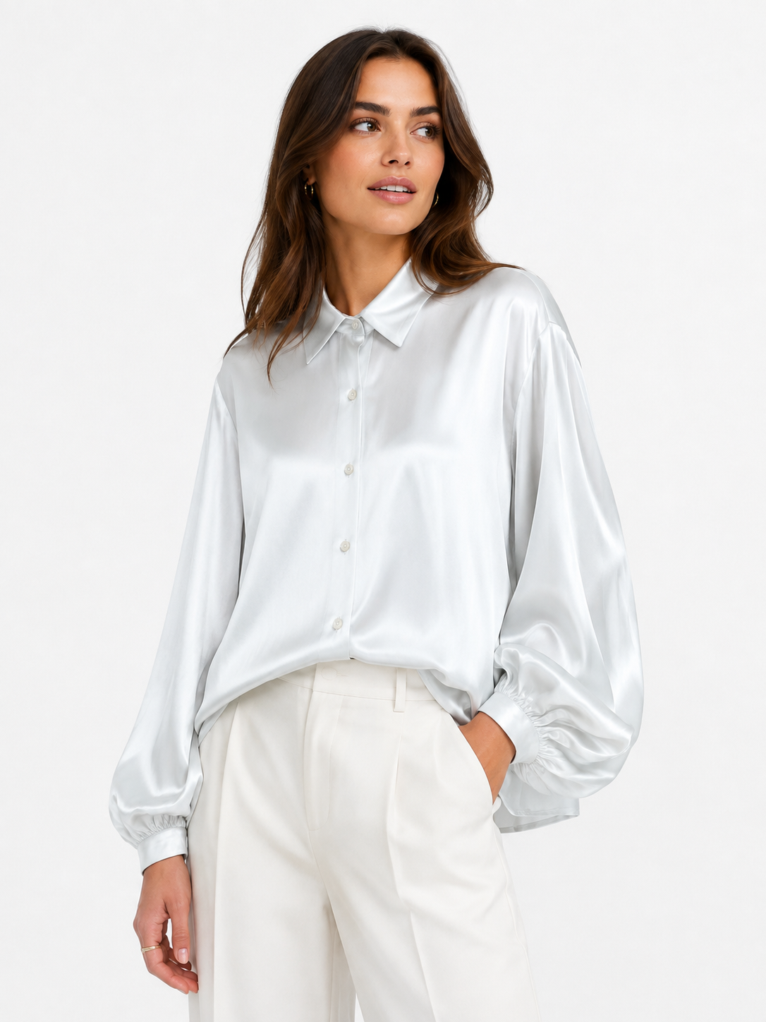 Eine Frau steht vor einem schlichten weißen Hintergrund, blickt zur Seite und trägt die Bluse „Antonia Shiny IceGrey“ im Oversize-Look mit Ballonärmeln sowie eine cremefarbene Hose.