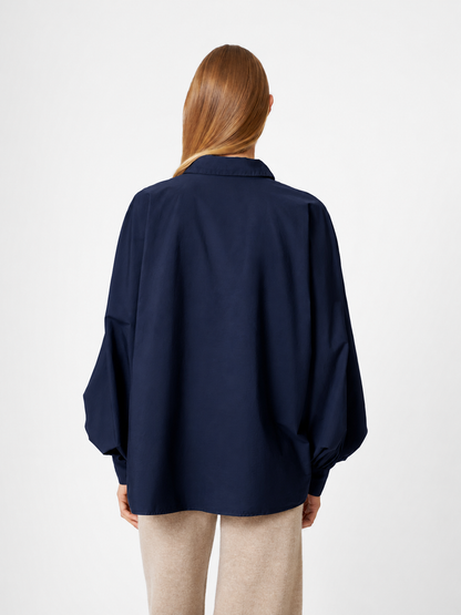 Eine Person mit langen, glatten Haaren ist von hinten zu sehen. Sie trägt eine locker sitzende Bluse „Antonia Cotton Navy One Size“ mit langen Ärmeln und eine hellbeige Hose vor einem schlichten weißen Hintergrund.