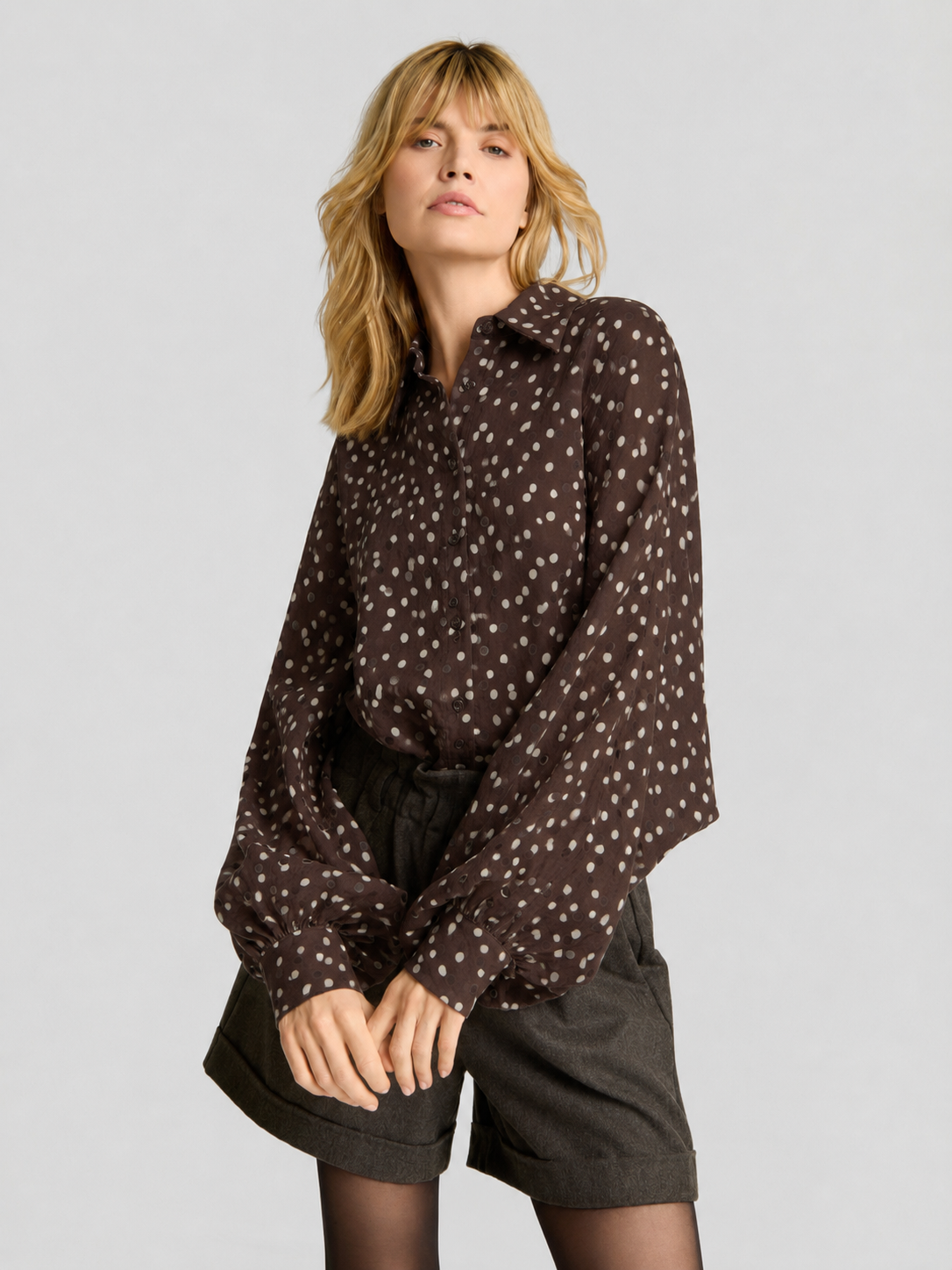 Eine Frau mit blonden Haaren trägt die Bluse „Antonia Polka Dots Brown Ecru“ zu dunklen Shorts. Vor einem schlichten hellgrauen Hintergrund steht sie in einer entspannten, selbstbewussten Pose und unterstreicht damit die mühelose Eleganz dieser in Italien hergestellten Damenbluse.