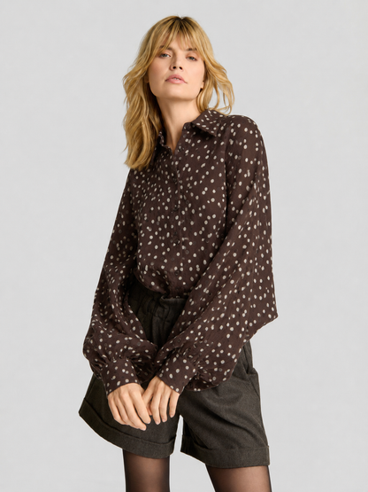 Eine Frau mit blonden Haaren trägt die Bluse „Antonia Polka Dots Brown Ecru“ zu dunklen Shorts. Vor einem schlichten hellgrauen Hintergrund steht sie in einer entspannten, selbstbewussten Pose und unterstreicht damit die mühelose Eleganz dieser in Italien hergestellten Damenbluse.