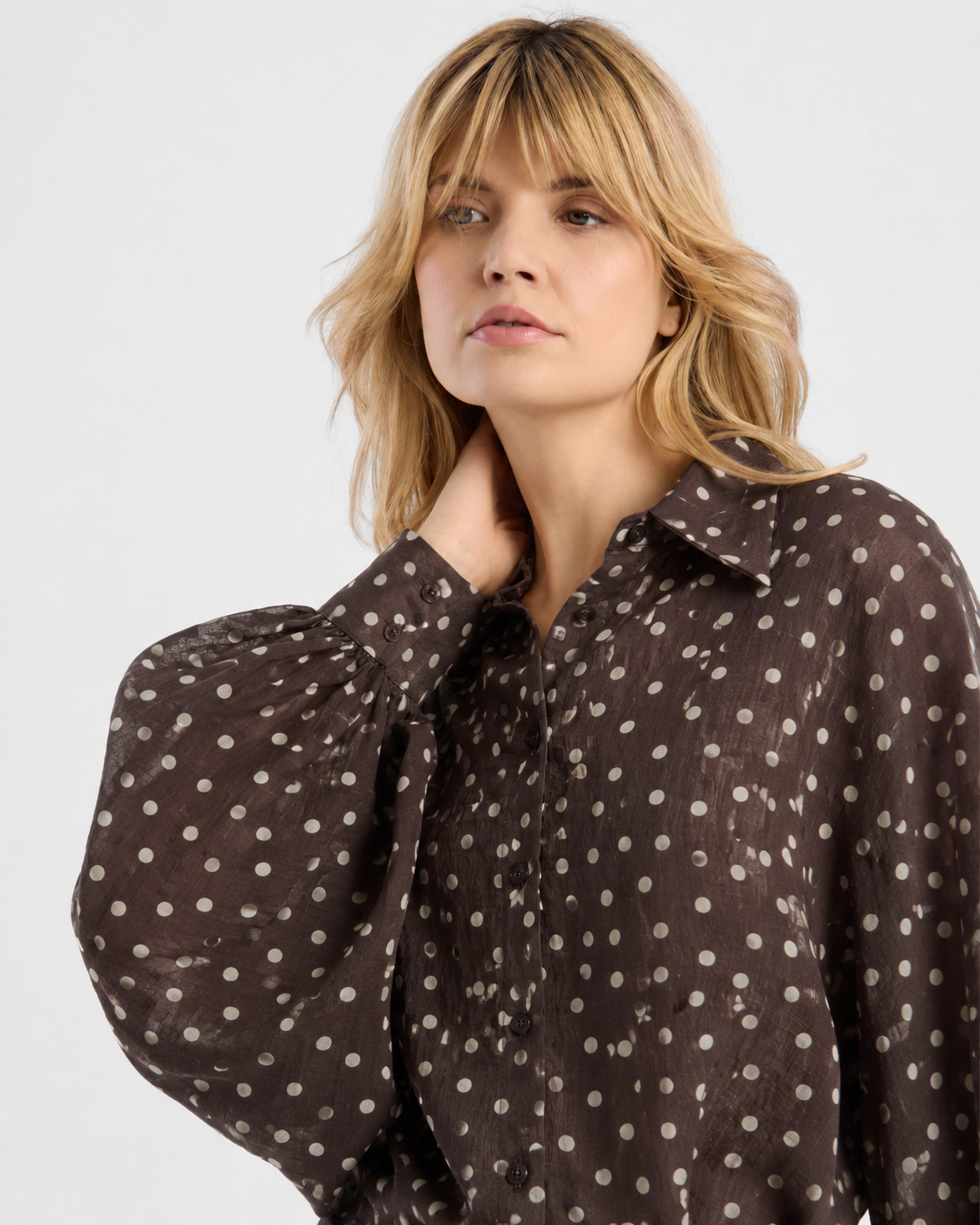 Eine Frau mit blonden Haaren trägt die Bluse „Antonia Polka Dots Brown Ecru“ – mit klassischen weißen Punkten – und steht vor einem schlichten Hintergrund, wobei sie sich an den Hals fasst und leicht zur Seite blickt. Diese stilvolle Damenbluse „Made in Italy“ verleiht ihrem zurückhaltenden Look einen Hauch von Eleganz.
