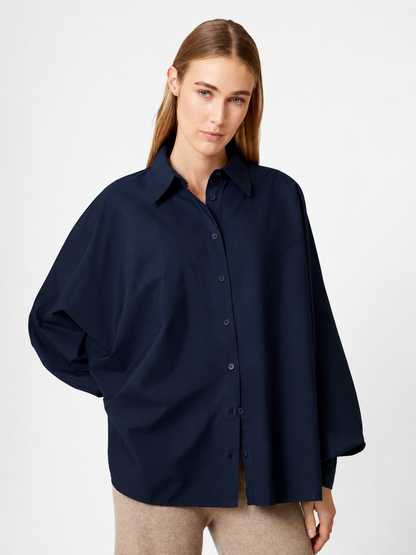 Eine Person mit langen, glatten, hellbraunen Haaren trägt eine übergroße Bluse „Antonia Cotton Navy“ (Einheitsgröße) und eine beige Hose und steht mit einer Hand hinter dem Rücken vor einem schlichten, hellen Hintergrund.