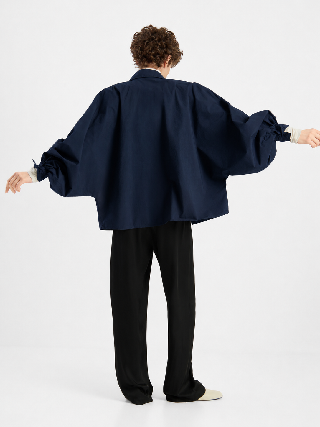 Eine Person steht mit dem Rücken zur Kamera, die Arme leicht ausgestreckt. Sie trägt die Bluse „Antonia Cotton Navy One Size“ – ein locker sitzendes, dunkelblaues Oberteil mit weiten Ärmeln –, dazu eine schwarze Hose und helle Schuhe. Der Hintergrund ist schlicht weiß.