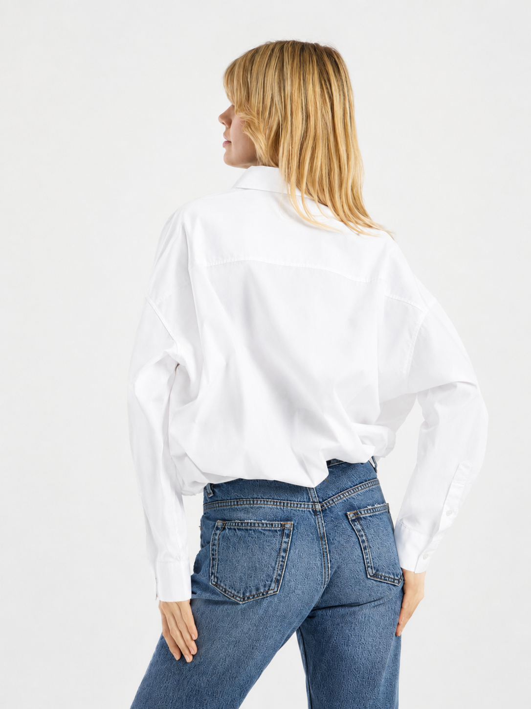Eine Person mit blonden Haaren, die eine locker sitzende weiße Ballonbluse aus Baumwolle und eine blaue Jeans trägt, steht mit dem Rücken zur Kamera, eine Hand in die Hüfte gestützt, vor einem schlichten Hintergrund.