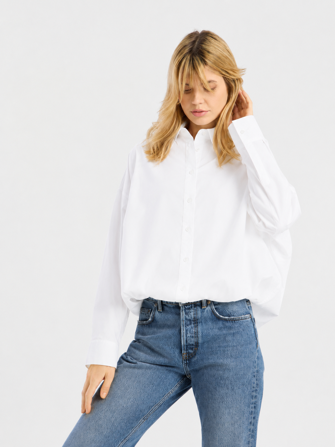 Eine Person, die eine locker sitzende, kurz geschnittene „Ballon Bluse“ in Weiß und eine blaue Jeans trägt, steht vor einem schlichten, hellen Hintergrund, blickt nach unten und berührt mit einer Hand ihr Haar.