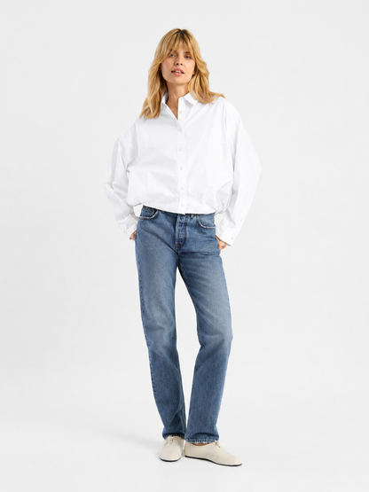 Vor einem weißen Hintergrund steht eine Frau, die die „Ballon Bluse White“ trägt, dazu blaue Jeans und weiße Schuhe. Sie hat blondes Haar, die Hände in die Vordertaschen gesteckt, und blickt mit einem sanften Lächeln leicht zur Seite.