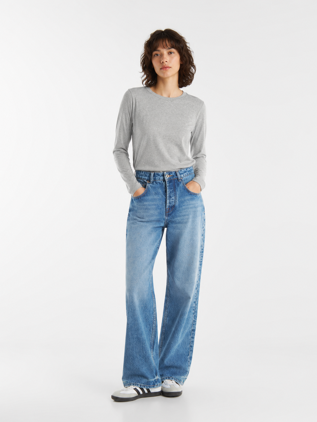 Eine Person mit schulterlangem braunem Haar trägt ein graues Langarmshirt, eine blaue Jeans mit weitem Bein und weiße Sneaker und steht mit den Händen in den Taschen vor einem schlichten weißen Hintergrund.