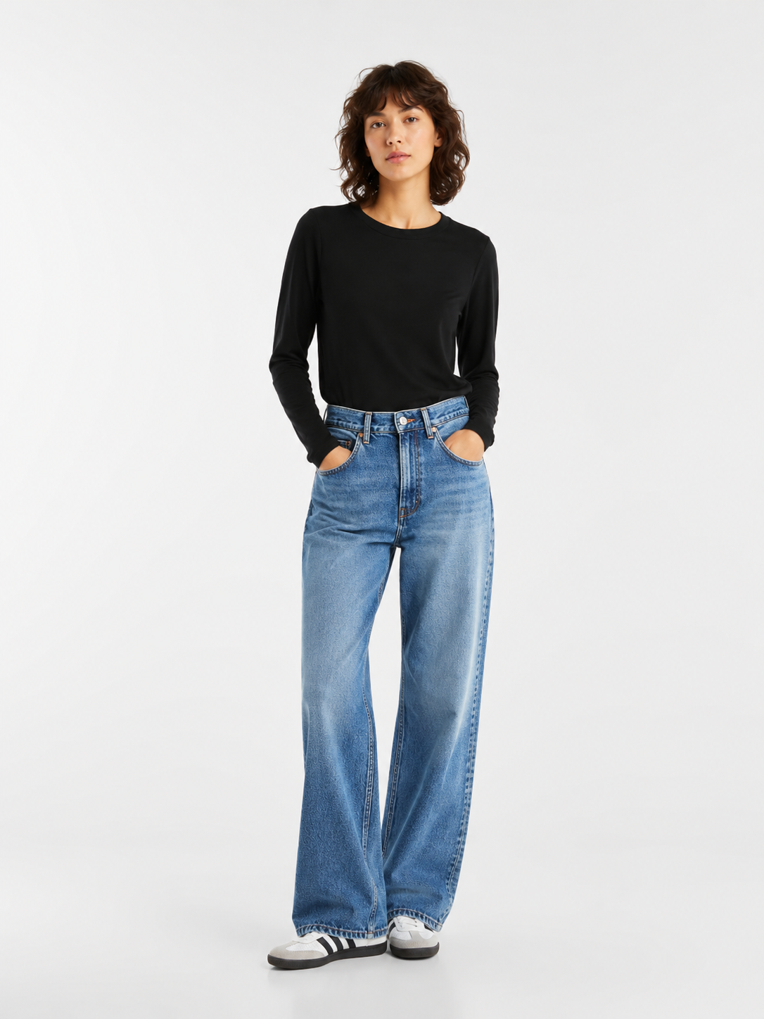 Eine Person, die mit den Händen in den Taschen vor einem schlichten weißen Hintergrund steht und ein schwarzes Langarmshirt, eine blaue Jeans mit weitem Bein sowie weiße Sneaker mit schwarzen Streifen trägt.