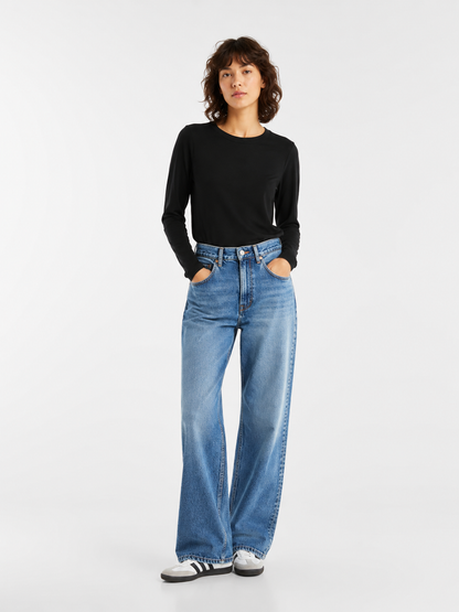 Eine Person, die mit den Händen in den Taschen vor einem schlichten weißen Hintergrund steht und ein schwarzes Langarmshirt, eine blaue Jeans mit weitem Bein sowie weiße Sneaker mit schwarzen Streifen trägt.