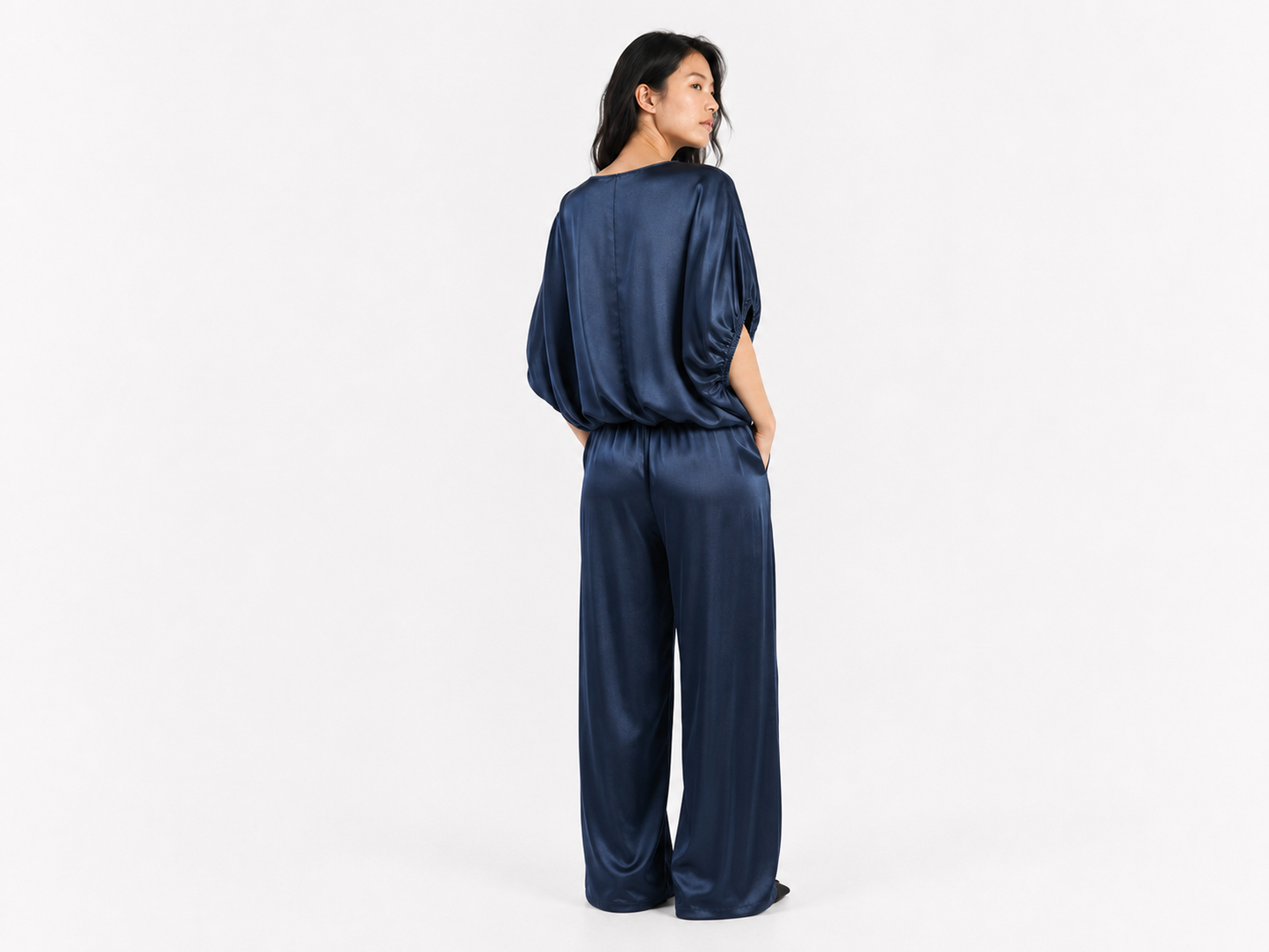 Eine Person steht mit dem Rücken zur Kamera und trägt die Bluse „Cowboy Shiny Navy“ aus Oversize-Satin mit stilvollen Fledermausärmeln sowie eine dazu passende Hose mit weitem Bein vor einem schlichten weißen Hintergrund.