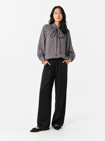 Eine Frau steht mit den Händen in den Taschen da und trägt die Bluse „Dotty Bow Grey Latte“ – eine graue Baumwoll-Voile-Bluse mit Polka-Dots und einer großen Schleife am Hals –, dazu eine schwarze Hose mit weitem Bein und schwarze Schuhe. Sie hat langes, welliges Haar und einen neutralen Gesichtsausdruck. Hergestellt in Italien.
