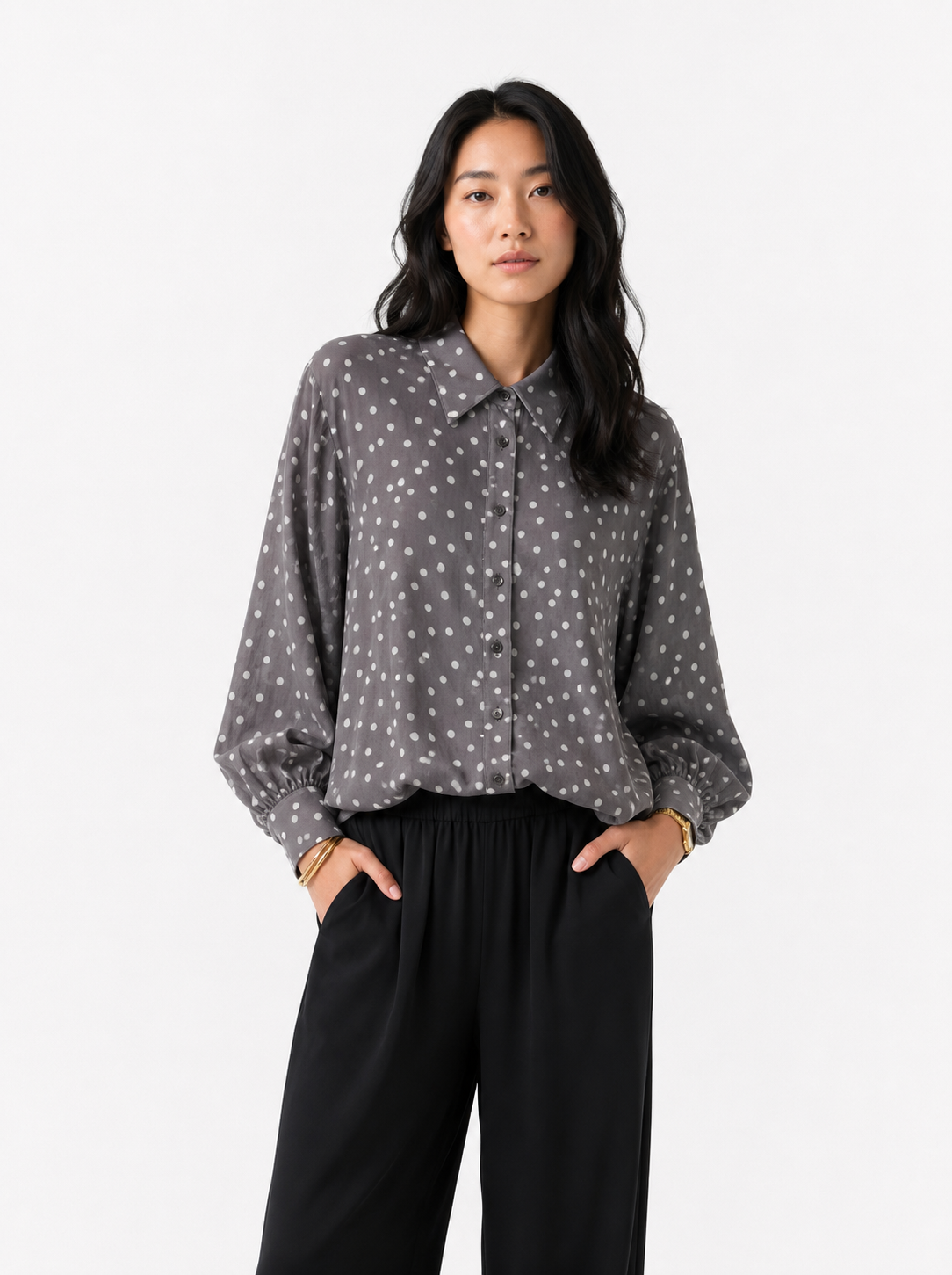 Eine Frau steht vor einem schlichten weißen Hintergrund, die Hände in den Taschen, und trägt eine schwarze Hose sowie die locker sitzende, langärmelige Bluse „Antonia Polka Dots Grey Latte“ – eine stilvolle Damenbluse aus Baumwoll-Voile in Grau mit weißen Punkten, die mühelos schick wirkt und in Italien hergestellt wurde.