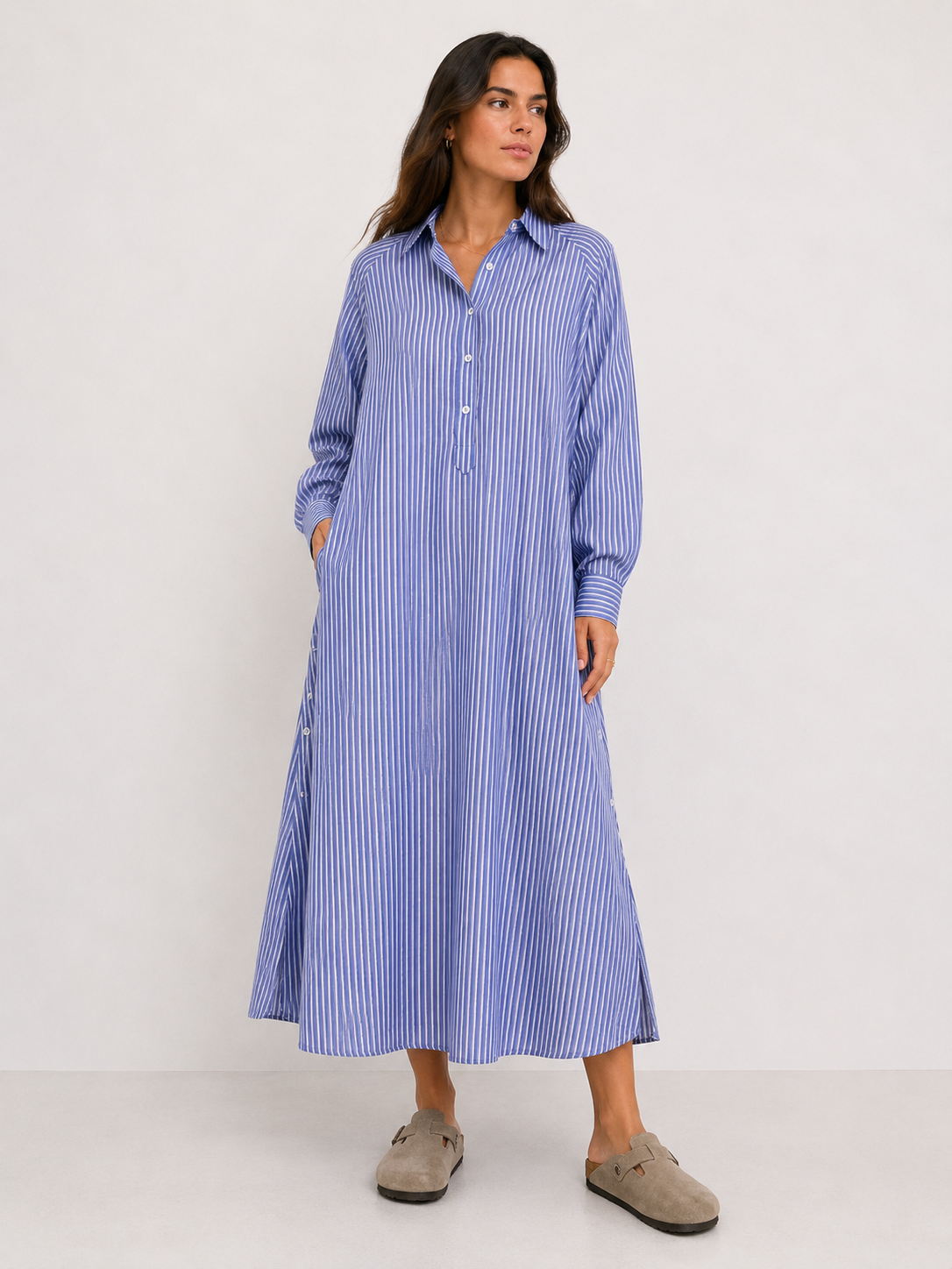 Eine Frau trägt das Kleid „Ocean“ in blau-weißen Streifen mit langen Ärmeln, einer Knopfleiste und Taschen. Sie kombiniert es mit beigen Slip-on-Schuhen und blickt leicht zur Seite.