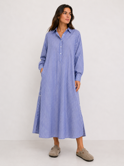 Eine Frau trägt das Kleid „Ocean“ in blau-weißen Streifen mit langen Ärmeln, einer Knopfleiste und Taschen. Sie kombiniert es mit beigen Slip-on-Schuhen und blickt leicht zur Seite.