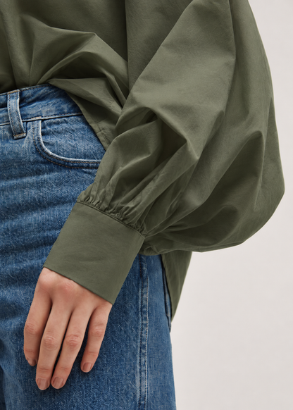 Eine Person, die blaue Jeans und ein olivgrünes, langärmeliges Hemd mit weit geschnittenen Ärmeln trägt, das an die stilvolle Bluse „Antonia Olive“ aus Baumwolle erinnert. Die linke Hand der Person ruht neben der Hüfte und unterstreicht die entspannte Eleganz dieser Bluse in Einheitsgröße.