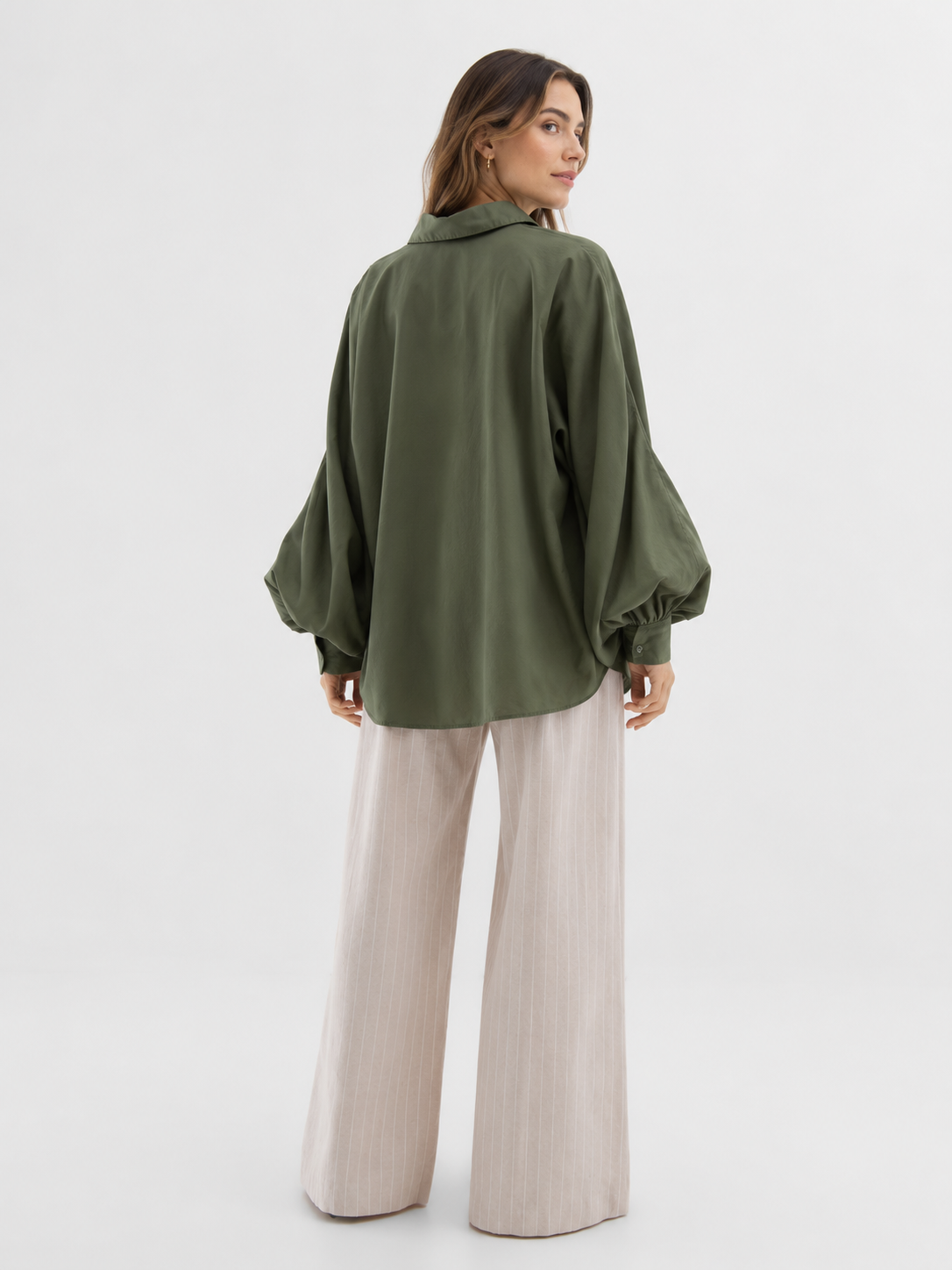 Eine Frau steht mit dem Rücken zur Kamera und trägt vor einem schlichten weißen Hintergrund die Bluse „Antonia Olive“ aus Baumwolle mit ausgestellten Ärmeln sowie eine beige Hose mit weitem Bein. Die Bluse in Einheitsgröße bietet eine lässige und zugleich elegante Silhouette.