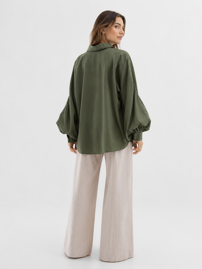 Eine Frau steht mit dem Rücken zur Kamera und trägt vor einem schlichten weißen Hintergrund die Bluse „Antonia Olive“ aus Baumwolle mit ausgestellten Ärmeln sowie eine beige Hose mit weitem Bein. Die Bluse in Einheitsgröße bietet eine lässige und zugleich elegante Silhouette.