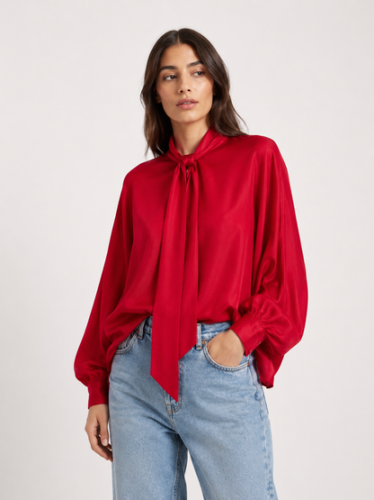 Eine Frau, die die Bluse „Antonia Bow Red“ mit einer Schleife am Hals und hellblaue Jeans trägt, steht vor einem schlichten weißen Hintergrund.