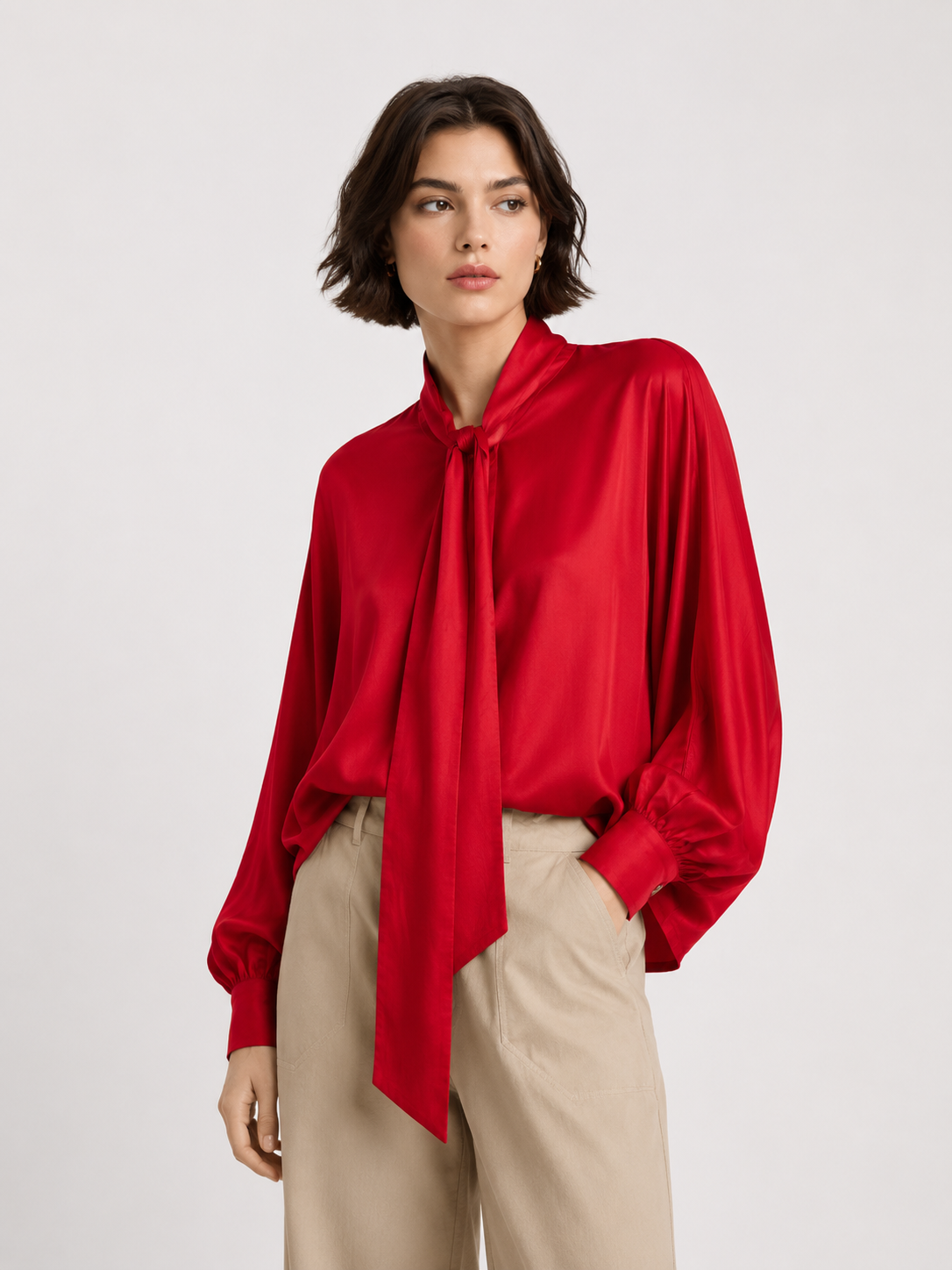 Eine Person mit kurzen braunen Haaren trägt die „Bluse Antonia Bow Red“, eine seidige Bluse mit einer langen Schleife, die an eine klassische Schleifenbluse erinnert. Dazu trägt sie eine beige Hose und steht vor einem schlichten, hellen Hintergrund.