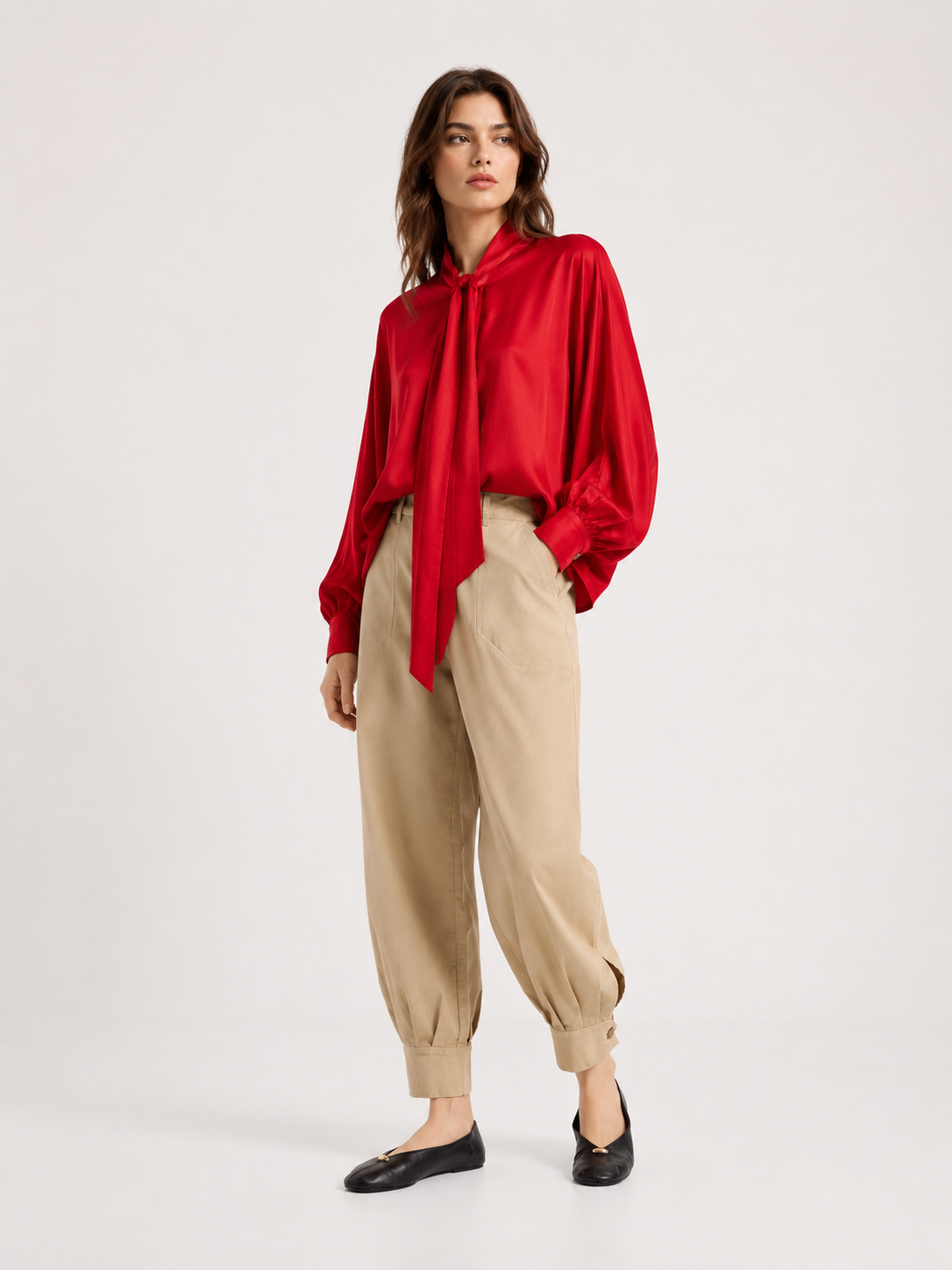 Eine Person steht vor einem schlichten, hellen Hintergrund und trägt die Bluse „Antonia Bow Red“ mit Bindeband am Hals, dazu eine beige Hose mit hoher Taille und schwarze Ballerinas.