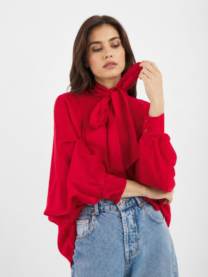 Eine Frau, die die Bluse „Antonia Bow Red“ und eine blaue Jeans trägt, schaut nach unten, während sie die Schleife am Hals festhält; der Hintergrund ist schlicht weiß.