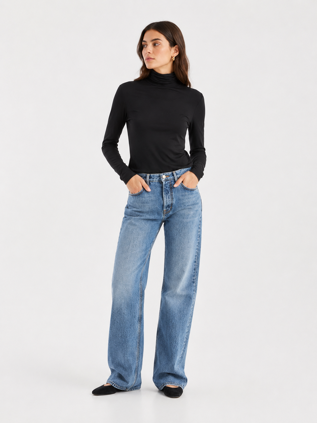 Eine Frau steht mit den Händen in den Taschen da und trägt das „Turtle Jersey Black“ – ein schwarzes Rollkragen-Shirt –, dazu hellblaue Jeans mit weitem Bein und schwarze Schuhe. Vor einem schlichten weißen Hintergrund blickt sie nach links und präsentiert einen zeitlosen Look, der in Portugal gefertigt wurde.