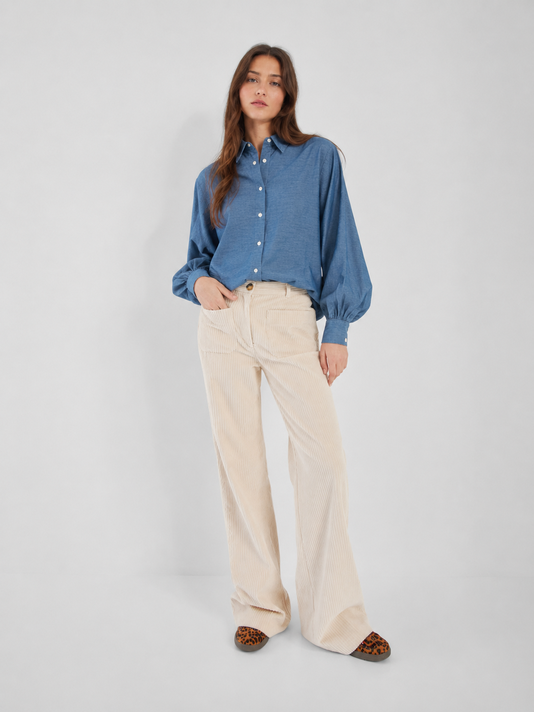 Eine Frau posiert vor einem schlichten, hellen Hintergrund und trägt die „Bluse Antonia Slim Denim“ – eine schicke Jeansbluse für Damen, hergestellt in Portugal –, kombiniert mit einer beigen Cordhose mit weitem Bein und flachen Schuhen mit Leopardenmuster.