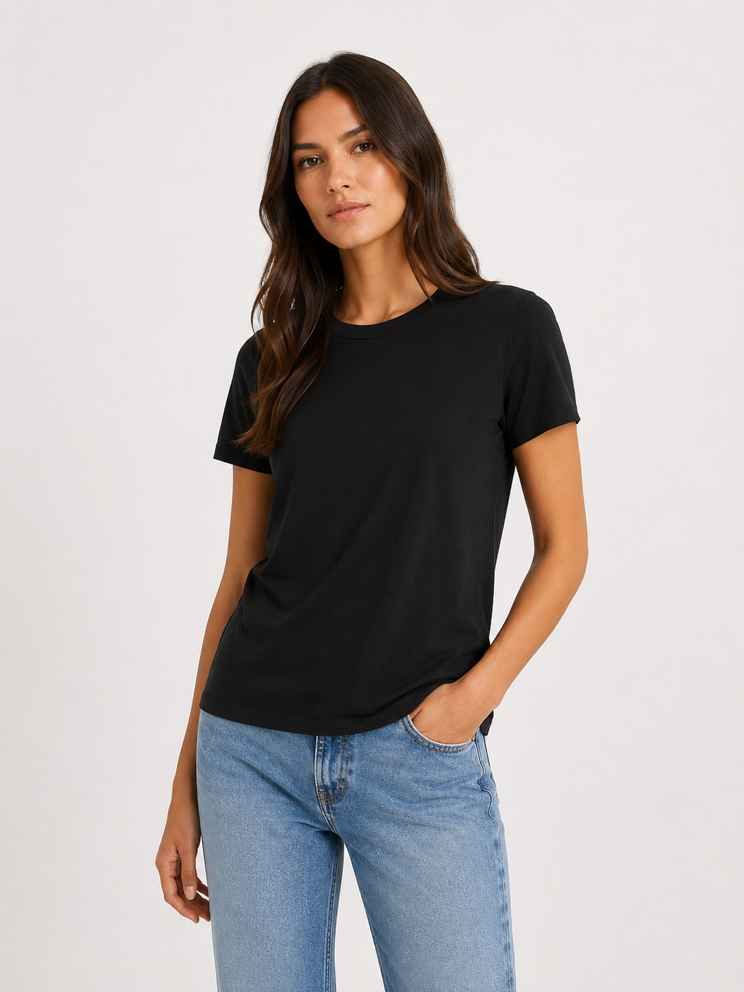 Eine Frau mit langen braunen Haaren, die ein schlichtes schwarzes T-Shirt mit Rundhalsausschnitt und eine hellblaue Jeans trägt, steht vor einem schlichten hellen Hintergrund und blickt mit neutralem Gesichtsausdruck in die Kamera. Eine Hand steckt in ihrer Hosentasche.