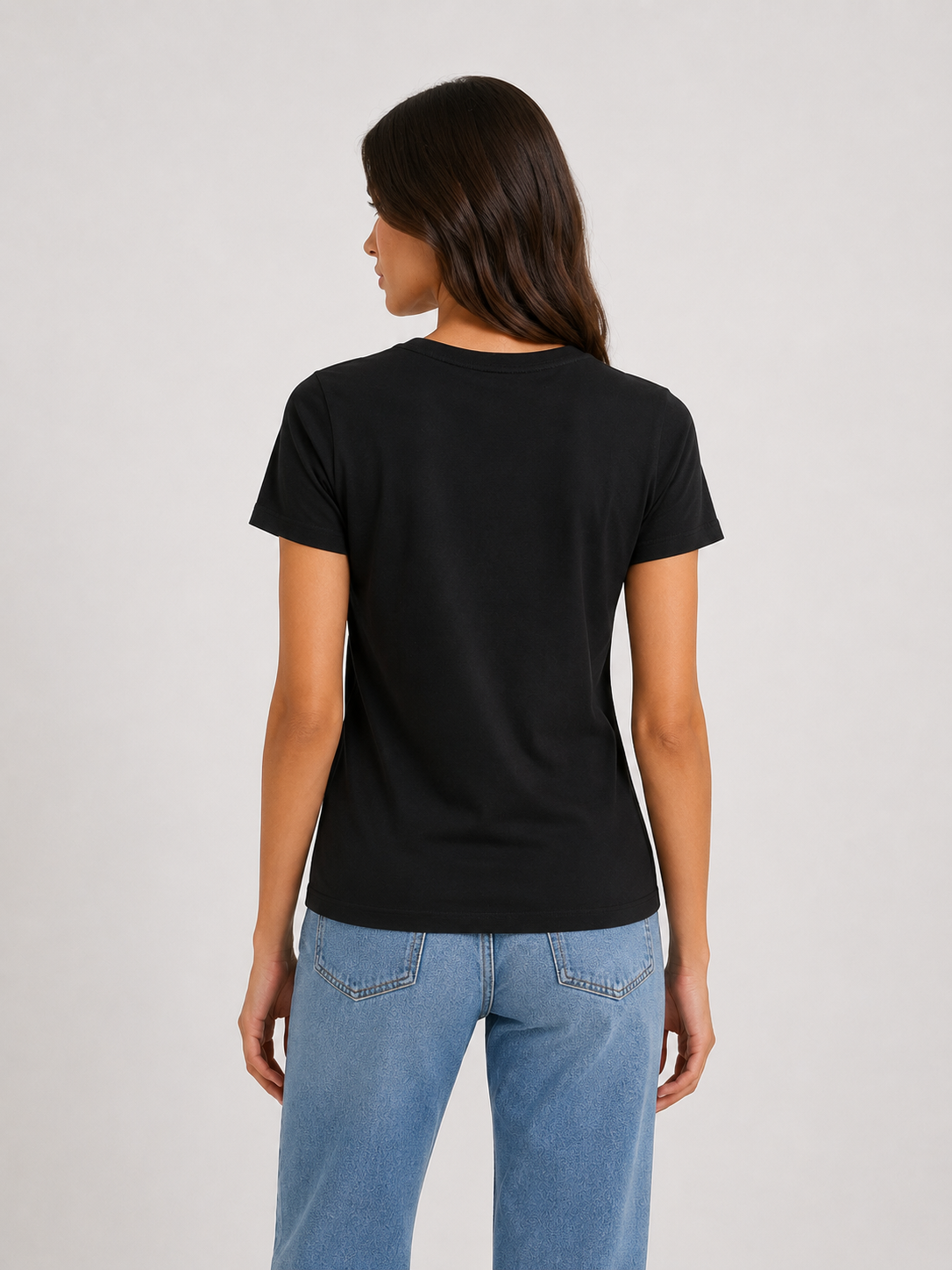 Eine Person mit langen, dunklen Haaren steht mit dem Rücken zur Kamera und trägt ein schwarzes T-Shirt mit Rundhalsausschnitt sowie hellblaue Jeans vor einem schlichten, hellen Hintergrund.