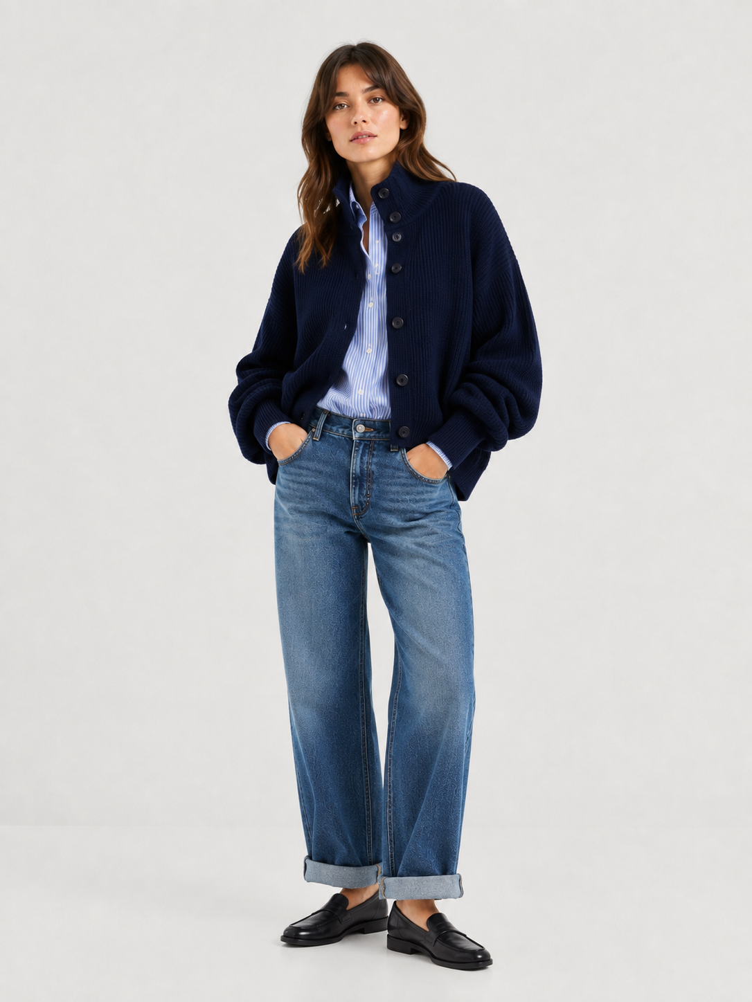 Vor einem schlichten, hellen Hintergrund steht eine Frau mit den Händen in den Taschen. Sie trägt die Strickjacke „Bomber Dunkelblau“ über einem blau gestreiften Hemd, eine Jeans mit weiten Bündchen und schwarze Slipper.