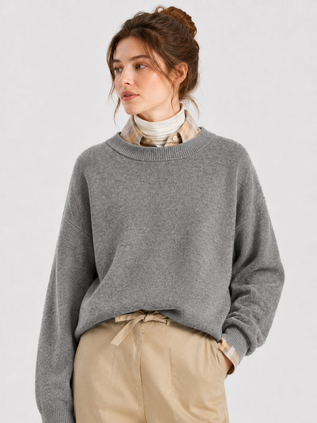 Eine Frau mit braunen Haaren, die zu einem Dutt zusammengebunden sind, trägt den Pullover „Giulia Grau“ über einem karierten Hemd und einem cremefarbenen Rollkragenpullover, dazu eine beige Hose; sie steht vor einem schlichten, hellen Hintergrund.