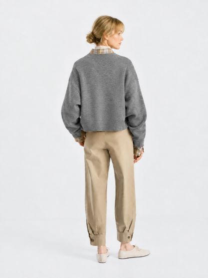 Eine Person steht mit dem Rücken zur Kamera und trägt den Pullover „Giulia Grau“ über einem karierten Hemd, eine beige Hose mit geknöpften Hosenbeinen und beige Schuhe vor einem schlichten, hellen Hintergrund.