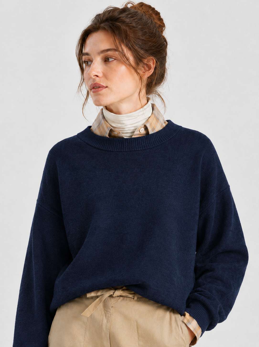 Eine Frau mit braunen Haaren, die zu einem Knoten zusammengebunden sind, trägt den Pullover „Giulia Dunkelblau“ aus Merinowolle über einem karierten Hemd und einer beigen Hose und blickt vor einem einfarbigen Hintergrund zur Seite.
