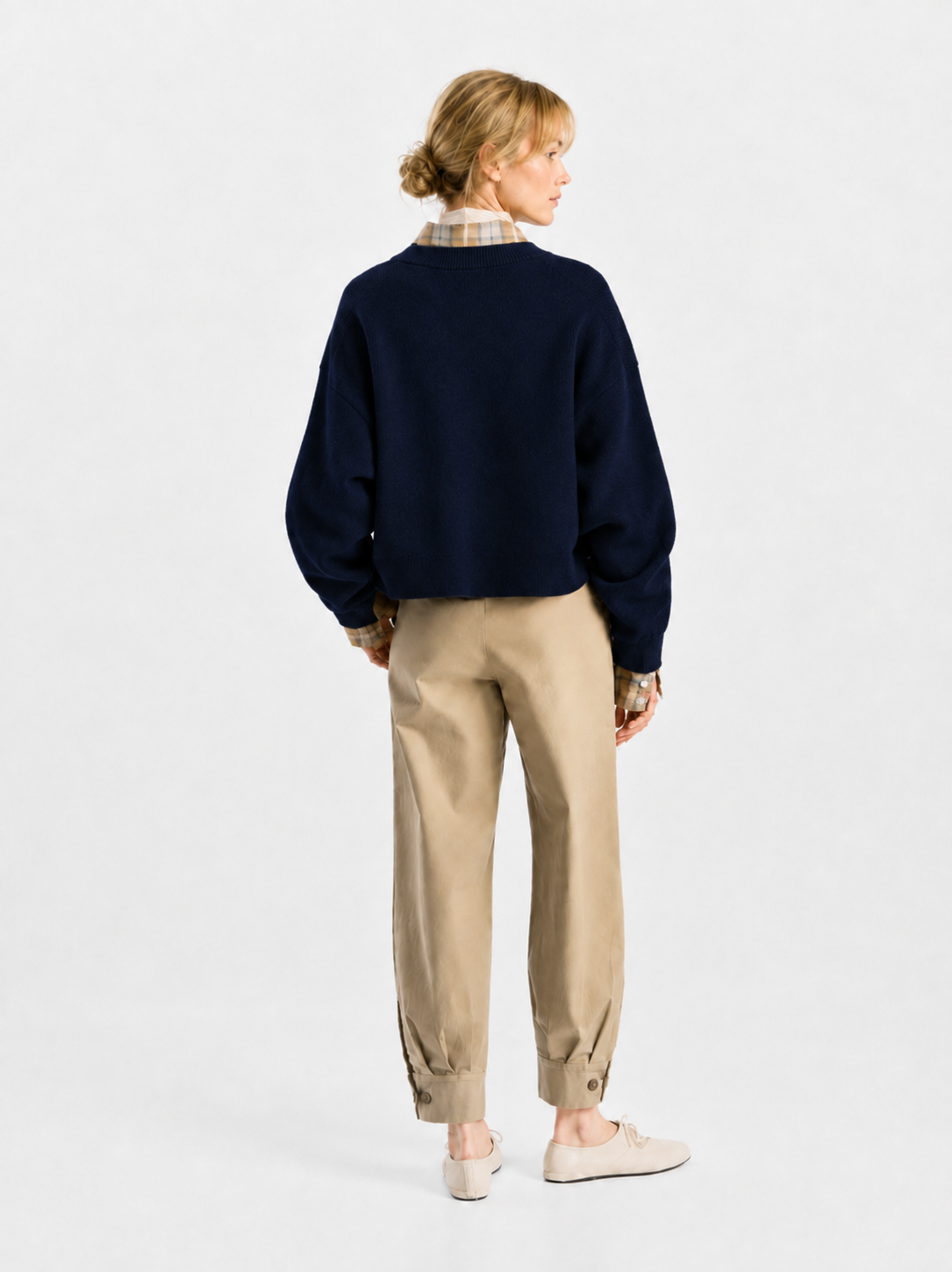 Eine Person steht mit dem Rücken zur Kamera und trägt den Pullover „Giulia Dunkelblau“ aus feiner Merinowolle über einem karierten Hemd, eine beige Hose mit Umschlag und Knopfdetails sowie cremefarbene Schuhe. Der Hintergrund ist schlicht und weiß.