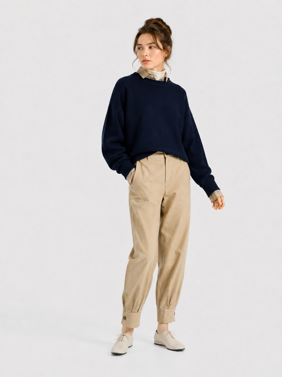 Eine Person steht vor einem schlichten Hintergrund und trägt den Pullover „Giulia Dunkelblau“ aus Merinowolle über einem Hemd mit Kragen, eine beige Plisseehose mit Umschlag sowie helle Schuhe. Die Haare sind hochgesteckt, und eine Hand steckt in der Hosentasche.