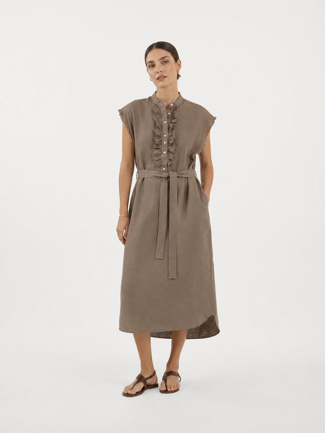 Eine Frau in einem ärmellosen, mit einem Gürtel versehenen Midikleid „Kleid Barcelona Soft Brown“ mit geknöpften Rüschen-Details auf der Vorderseite, die vor einem schlichten weißen Hintergrund steht und braune Sandalen trägt.