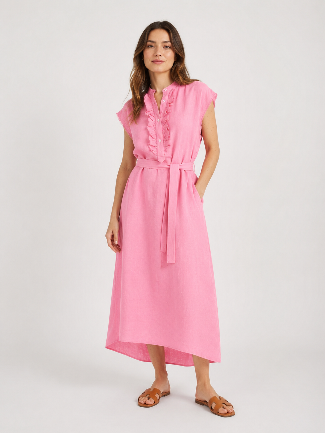 Vor einem schlichten weißen Hintergrund steht eine Frau, die das ärmellose, knöchellange Kleid „Barcelona Pink“ trägt, das sich durch eine Rüschenverzierung auf der Vorderseite und einen Bindegürtel in der Taille auszeichnet, dazu trägt sie braune Sandalen.