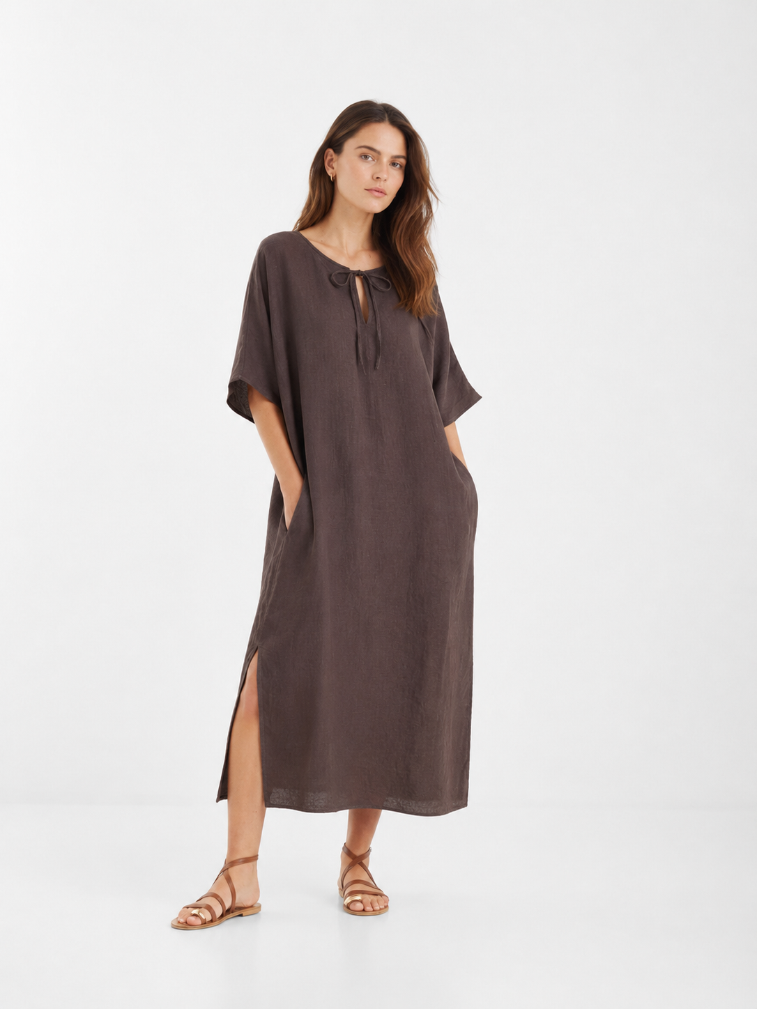 Eine Frau trägt das „Dress Kaftan Linen Brown“ – ein locker sitzendes, braunes Maxikleid aus Leinen mit kurzen Ärmeln, Seitenschlitzen und einem Bindegürtel am Ausschnitt. Sie steht mit den Händen in den Taschen des Kleides und kombiniert es mit flachen, braunen Sandalen vor einem schlichten weißen Hintergrund. Dieses nachhaltige Maxikleid aus Leinen vereint stilvollen Komfort mit umweltbewusstem Design.