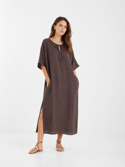 Eine Frau trägt das „Dress Kaftan Linen Brown“ – ein locker sitzendes, braunes Maxikleid aus Leinen mit kurzen Ärmeln, Seitenschlitzen und einem Bindegürtel am Ausschnitt. Sie steht mit den Händen in den Taschen des Kleides und kombiniert es mit flachen, braunen Sandalen vor einem schlichten weißen Hintergrund. Dieses nachhaltige Maxikleid aus Leinen vereint stilvollen Komfort mit umweltbewusstem Design.