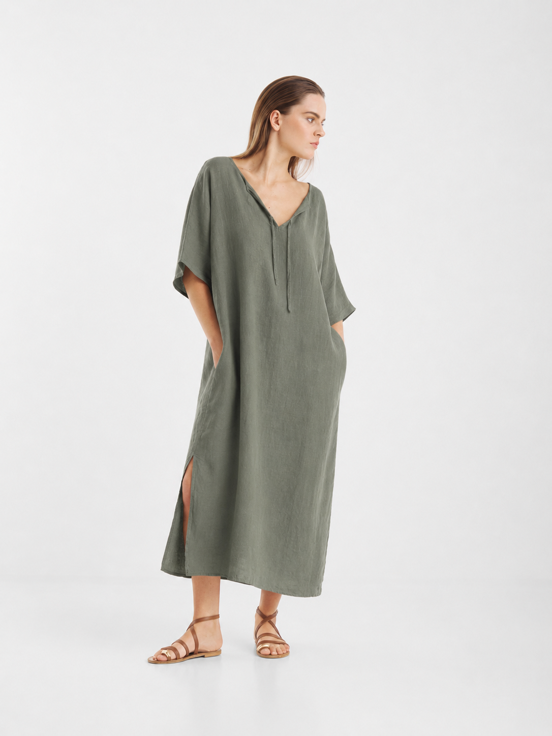 Eine Frau trägt einen locker sitzenden, olivgrünen Kaftan aus Leinen („Dress Kaftan Linen Olive“) mit kurzen Ärmeln, V-Ausschnitt und Seitenschlitzen. Sie trägt hellbraune flache Sandalen und hat die Hände in den Taschen des Kleides. Der nachhaltig hergestellte Kaftan besticht durch eine bequeme Passform und eignet sich daher ideal für den Alltag. Der Hintergrund ist schlicht weiß.