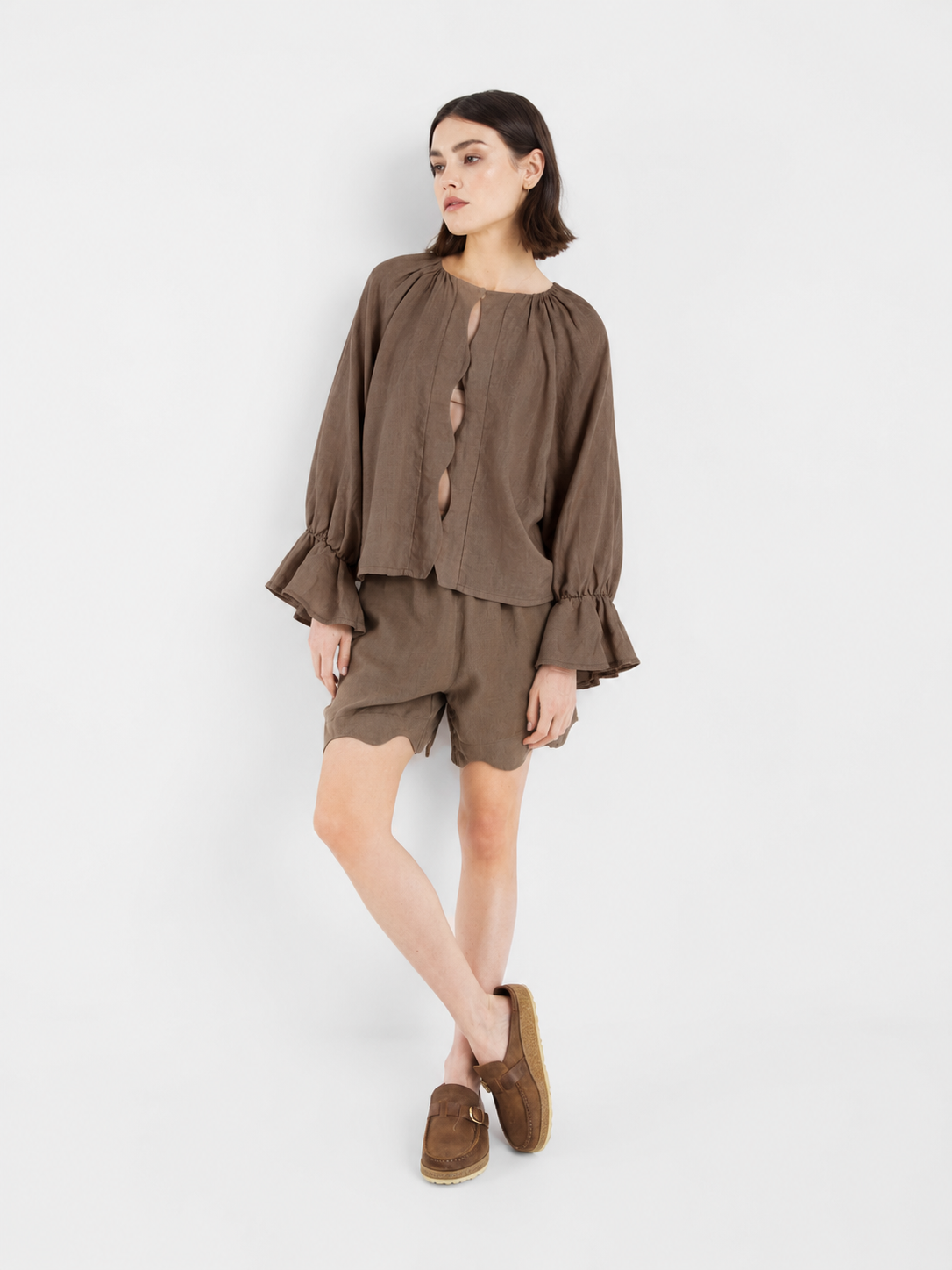 Vor einem weißen Hintergrund steht eine Frau, die die locker sitzende Bluse „Wave Linen Soft Brown“ mit gerüschten Ärmelbündchen, dazu passende braune Shorts mit Wellenkanten und braune Slipper trägt. Sie blickt zur Seite, wobei ein Bein leicht angewinkelt ist, und verkörpert damit einen entspannten Sommerlook.