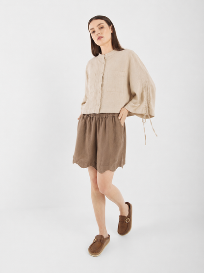 Eine Frau in einer beigen Bluse „Mary Linen Beige“ mit Ballonärmeln und braunen Shorts steht vor einem schlichten weißen Hintergrund, die Hände in den Taschen, und trägt braune Slipper. Ihre Bluse „Mary Linen Beige“ verfügt über verstellbare Ärmel, die für zusätzliche Vielseitigkeit und Stil sorgen.