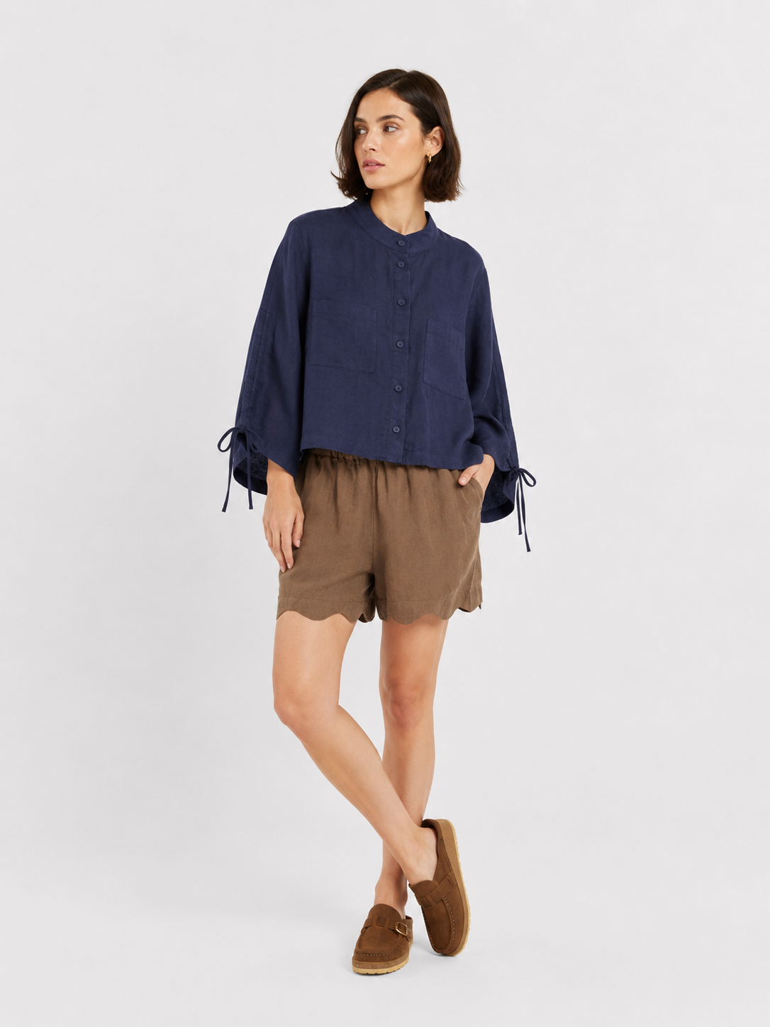 Vor einem weißen Hintergrund steht eine Frau, die die Bluse „Mary Linen Navy“ mit verstellbaren Ärmeln und Schleifendetails trägt, dazu braune Shorts mit Wellenrand und braune Slipper. Sie blickt zur Seite und hat eine Hand in der Tasche.