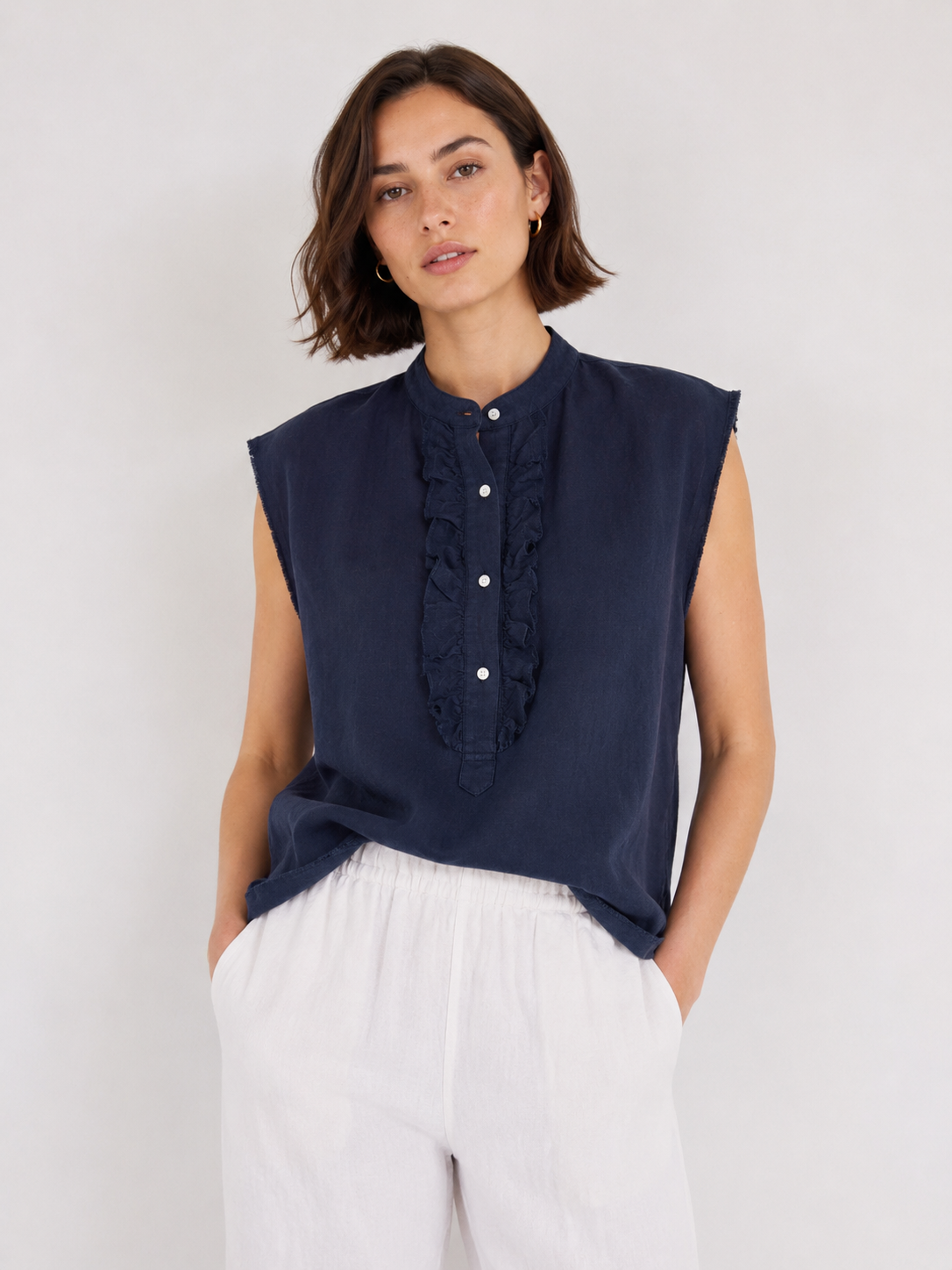 Eine Frau mit kurzen braunen Haaren trägt die ärmellose, marineblaue Bluse „Barcelona Linen“ mit zarten Rüschendetails, dazu eine weiße Hose. Sie steht vor einem schlichten, hellen Hintergrund und hat die Hände in den Taschen.