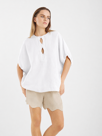 Vor einem schlichten, hellen Hintergrund steht eine Frau mit einer Hand in der Tasche. Sie trägt die Bluse „Cowboy Wave Linen White“ – eine locker sitzende weiße Leinenbluse mit Schlüssellochausschnitt – kombiniert mit beigen Shorts mit Wellenrand.