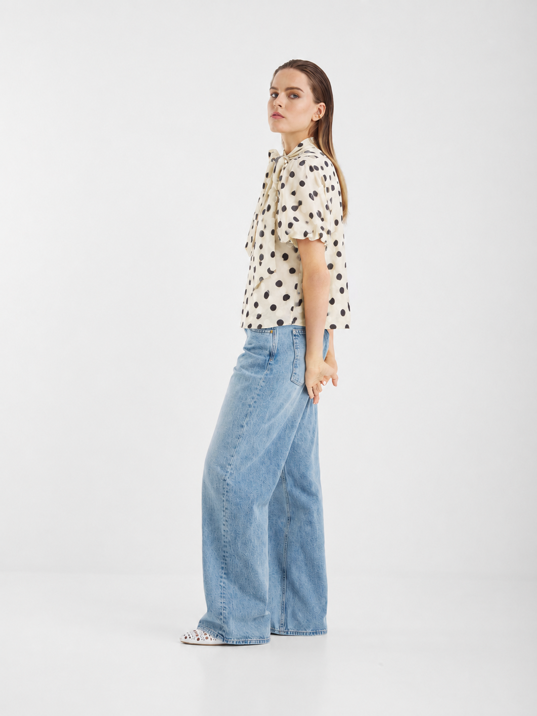 Vor einem schlichten weißen Hintergrund steht eine Person in seitlicher Haltung und trägt eine „Bluse Bow Dots Ecru Black“ – eine Bluse aus ecrufarbenem Baumwollvoile mit verspielten schwarzen Punkten – sowie hellblaue Jeans mit weitem Bein.