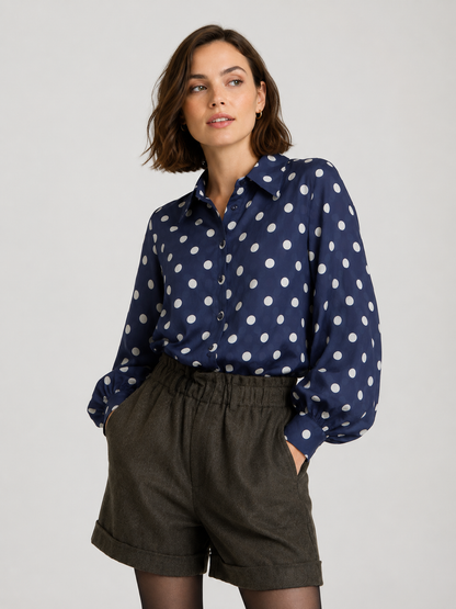 Eine Frau steht vor einem schlichten, hellen Hintergrund, die Hände in den Taschen ihrer grauen Shorts mit hoher Taille, und trägt die Oversize-Bluse „Antonia Dots Navy Ecru“ – gefertigt aus luftigem Baumwoll-Voile mit einem marineblau-weißen Polka-Dot-Muster. Hergestellt in Italien.